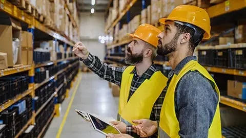Two men in a warehouse looking at shelves, wearing orange hard hats and yellow safety vests