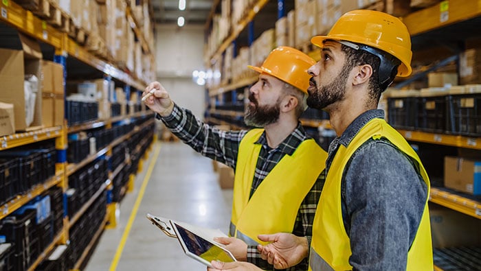 Two men in a warehouse looking at shelves, wearing orange hard hats and yellow safety vests